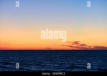 Tiefblaue Ozeanwellen unter verlaufendem Himmel, die von hellorange zu hellblau übergehen, mit verstreuten Wolken nahe der Horizontlinie und ruhigem geldautomaten Stockfoto
