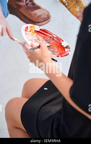 Maler, der die Farbpalette mit hellroten und gelben Acrylfarben hält und bei Tageslicht mit feinen Pinseln Striche aufträgt. Das zwanglose Ambiente konzentriert sich auf han Stockfoto
