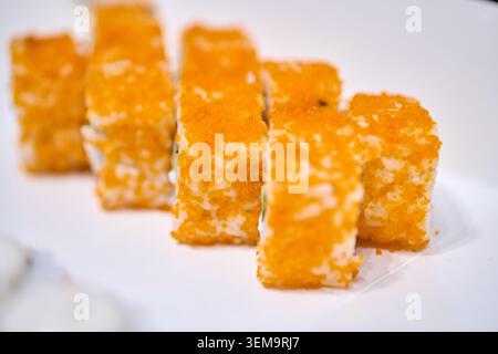 Mehrere Makizushi-Sushi-Brötchen, die mit leuchtendem orangenem Fischrogen beschichtet sind, werden ordentlich auf einer sauberen weißen Platte mit weicher natürlicher Beleuchtung platziert Stockfoto