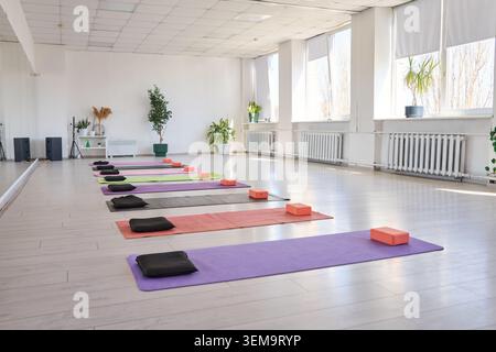 Sonnendurchflutete Yoga-Studio-Innenausstattung mit gleichmäßig verteilten Matten in verschiedenen Farben mit schwarzen Kissen und orangefarbenen Blöcken in einem sauberen, weißen Raum mit Pflanzen und Stockfoto