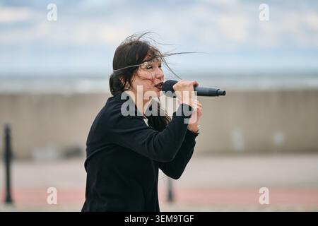 Fokussierte junge Erwachsene Frau, die in ein Mikrofon singt, in schwarzem Cosplay-Outfit im Freien mit bewölktem Himmel und verschwommenem Hintergrund. Wind weht Stockfoto