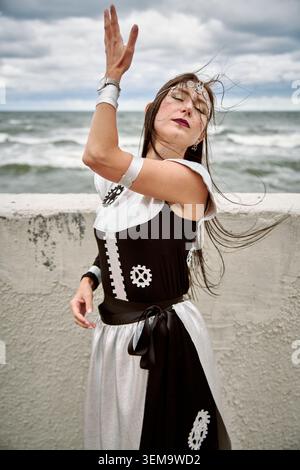 Erwachsene Frau mit langen dunklen Haaren in schwarz-weißem Cosplay-Kleid mit Gear-Mustern posiert anmutig am Ozean unter stimmungsvollem bewölktem Himmel, Wind weht Stockfoto