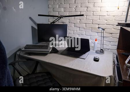 Kleiner Arbeitsbereich mit Laptop und Monitor auf überdachtem Tisch vor weißer Ziegelwand, beleuchtet durch Schreibtischlampe und Deckenleuchte bei niedrigen Umgebungstemperaturen Stockfoto