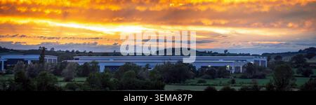 Ein moderner Industrielager-Komplex befindet sich in einer grünen ländlichen Landschaft unter einem dramatischen orangen Sonnenuntergang und schafft einen beeindruckenden Kontrast zwischen manuellem Hintergrund Stockfoto