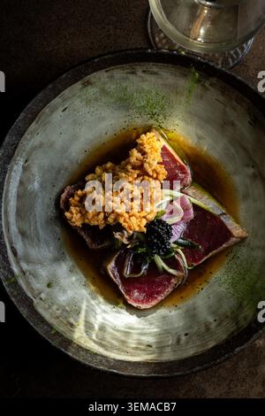 Eine Nahaufnahme leicht angebratener Thunfisch-Tataki-Scheiben, serviert mit schwarzem Kaviar, Mikrogrün, einer herzhaften Sauce und einem goldknusprigen Topping in einer dunklen cera Stockfoto