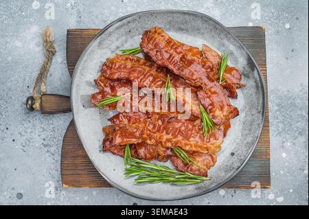 Goldene, brutzelnde Speckstreifen, die mit Rosmarin auf einem strukturierten Teller über hellem Betonhintergrund angeordnet sind, ideal für kulinarische Gestaltung. Stockfoto