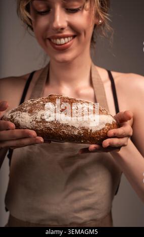 Lächelnde Frau, die eine Schürze trägt und einen frisch gebackenen Sauerteigbrot mit einer knusprigen Textur präsentiert Stockfoto