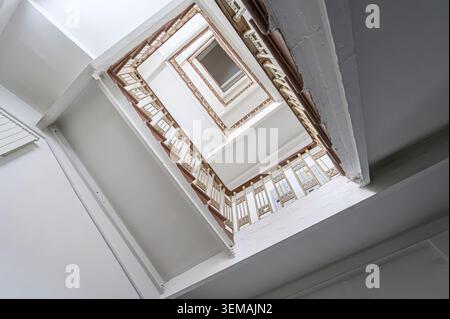 Niedrigwinkelaufnahme einer Treppe, mit Blick nach oben. Es hat weiße Decken und hellbraune Geländer, die ein quadratisches Spiralmuster zu einem Fenster bilden. Stockfoto