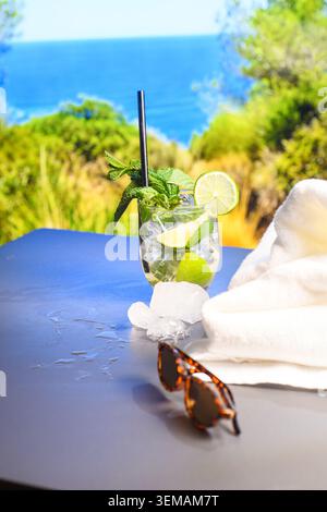 Eiskaltes Limetten- und Minzgetränk mit schwarzem Stroh neben einem weißen Handtuch und einer Sonnenbrille, fotografiert auf einer sonnigen mediterranen Terrasse am Meer. Stockfoto