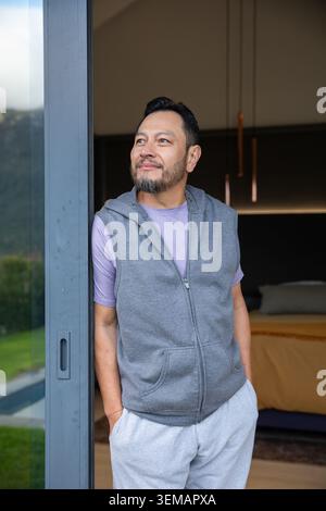 Reifer Erwachsener asiatischer Mann, der in einer Glasschiebetür steht, einen Hoodie trägt und auf den Pool blickt Stockfoto
