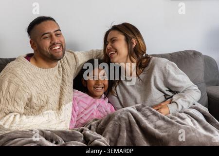 Eltern mit Kind sitzen zu Hause auf einer grauen Couch und teilen sich eine kuschelige Decke, während sie einen rosafarbenen Bademantel tragen Stockfoto