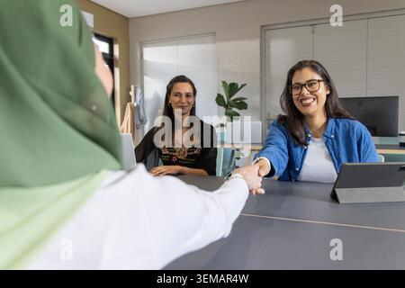 Erwachsene Frau mit blauem Hemd und Brille, die am Bürotisch mit Tablet die Hände schüttelt Stockfoto