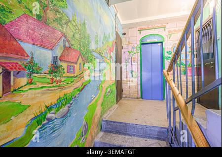 Ein Eingangsbereich mit einem farbenfrohen Wandbild von Häusern und einem Bach an der linken Wand. Eine blaue Fahrstuhltür und eine gemalte Ziegelwand rechts, zusammen mit einer Stockfoto