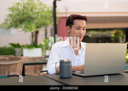 Ein landschaftliches Foto von einem nicht binären, geschlechtsabhängigen schwulen Thai-Mann aus Isan, der ferngesteuert an einem silbernen Laptop arbeitet. Stockfoto