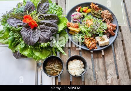 Gesunde vegetarische Bio-Mahlzeit mit frischem Gemüse, essbaren Blumen, gewürztem Tofu und Beilagen auf einem Holztisch sowie Salatvorhang zum Selbstbedienen. Stockfoto