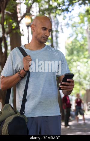 Afrikanischer Mann, der auf schattigem Weg steht, mit Telefon und schnappendem Sportgurt, mit grauem T-Stück Stockfoto