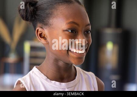 Afrikanische junge Frau lächelt, sieht nach rechts zu Hause, trägt eine helle Decke in der Nähe einer verschwommenen Pflanze Stockfoto