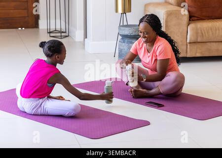 Afroamerikanische Mutter und Kind sitzen zu Hause auf lila Yogamatten in Aktivkleidung und einer Flasche Stockfoto