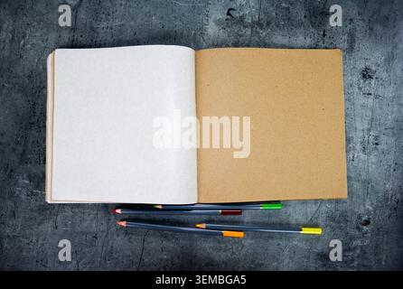 Ansicht von oben auf offenes Kraftpapier-Notizbuch mit Bleistiften auf grauem Steinhintergrund. Stockfoto
