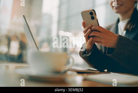 Lächelnde weiße junge Erwachsene Frau mit Smartphone neben Laptop in einem Café Stockfoto