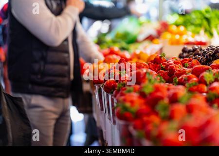 Frische rote Erdbeeren in Kunststoffbehältern auf der lokalen Markthalle in Riga Lettland, männlicher Kunde, der Bio-süße Su kauft Stockfoto
