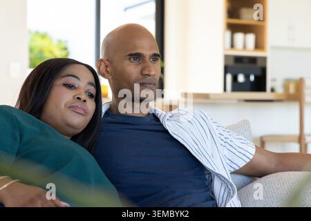 Ein Paar Afroamerikaner, ein indischer Mann, der zu Hause auf dem Sofa sitzt, sich beugt und ein gestreiftes Hemd trägt Stockfoto