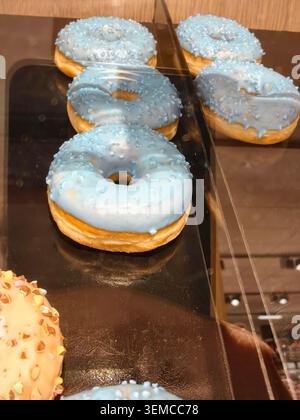 Eine Reihe glasierter Donuts mit hellblauer Zuckerglasur und Streuseln befindet sich auf einem Glasvitrine in einer Bäckerei. Die Donuts liegen auf einem dunklen Tablett mit warmer Beleuchtung Stockfoto