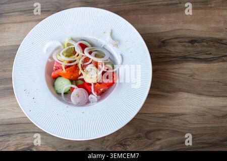 Scharfer, detaillierter Blick von oben auf einen frischen Gemüsesalat mit Tomaten, Gurken, Paprika und Zwiebeln, serviert in einer weißen Keramikschale auf einem hölzernen Ta Stockfoto