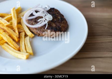 Scharfe, detaillierte Ansicht eines gegrillten Rindfleischschnitts mit geschnittenen Zwiebelringen, serviert mit goldenen Pommes frites auf einem weißen Teller, Referenz Stockfoto