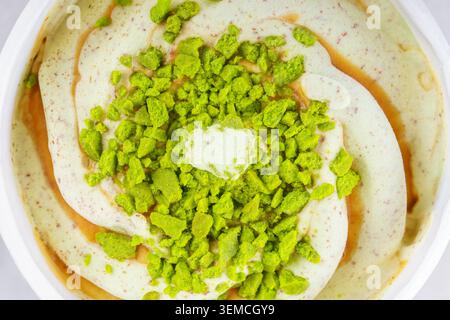 Pistazieneis in Papiereimer oder Becher mit Karamellsauce und zerstoßenen Pistazien. Draufsicht, Nahaufnahme, Makro. Dessert, Süßigkeiten, Dessert, Stockfoto