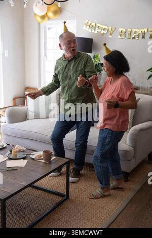 Asiatisches Paar tanzt und singt im Wohnzimmer, Senior trägt Kegelmütze unter HAPPY BIRTH Banner Stockfoto