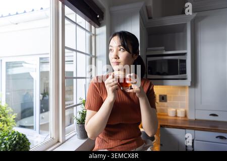 Asiatische Frau, die eine Teetasse aus Glas neben einer Topfpflanze hält, am Küchenfenster, mit Blick nach draußen, Kopierraum Stockfoto