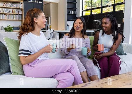 Drei Freundinnen sitzen im Wohnbereich auf dem Sofa, tragen Loungewear, halten Tassen und Smartphone Stockfoto
