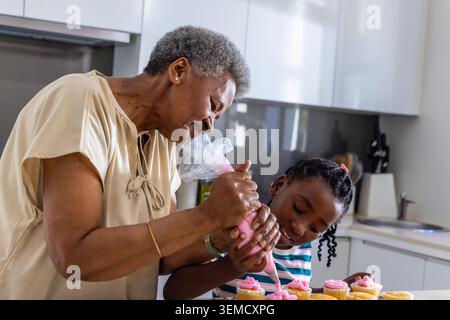 Ältere afrikanische Frau, die das afrikanische Jugendmädchen leitet, pinkfarbene Zuckerguss mit einem Paspelbeutel in der Küche Stockfoto