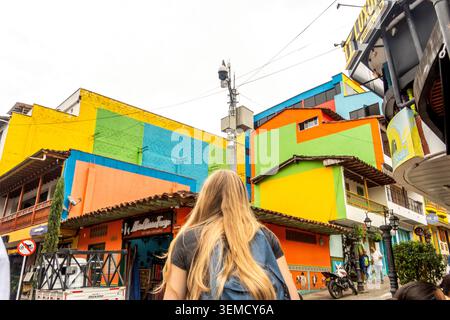 Eine Frau mit langen blonden Haaren, die von hinten gesehen wird, läuft eine lebhafte Straße entlang, die von farbenfrohen Wänden, Ladenfronten und urbaner Architektur unter d flankiert wird Stockfoto