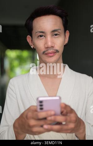 Ein vertikaler Nahporträt eines nicht-binären schwulen Mannes von Isan, thailändischer Abstammung, der direkt in das Kameraobjektiv blickt, mit einem ruhigen, selbstbewussten Blick Stockfoto