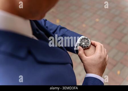 Ein Mann in einem eleganten blauen Anzug überprüft die Zeit auf seiner modernen Armbanduhr. Die Aufnahme vermittelt das Konzept von Vorausschau, Pünktlichkeit und Zeitmanagement. Stockfoto