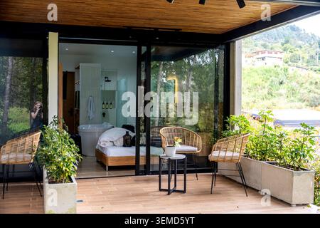 Eine moderne Außenterrasse verfügt über Korbstühle und einen kleinen Tisch, der vor großen Glastüren steht, die eine Schlafzimmersuite mit Topfpflanzen im Inneren freigeben Stockfoto