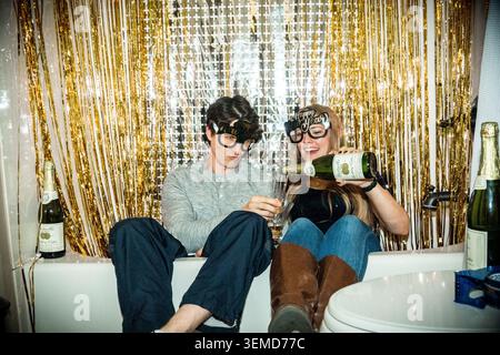 Zwei junge Menschen mit neuartigen Gläsern sitzen in einer weißen Badewanne, umgeben von goldenen Lametta-Vorhängen und Champagnerflaschen am Rand, was zu einem festi führt Stockfoto