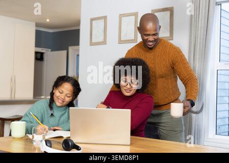 Eine vielfältige Familie sitzt am Holztisch, eine Frau, die am Laptop arbeitet, während das Kind mit Bleistift schreibt Stockfoto