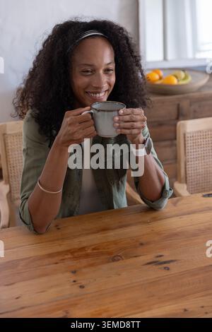 Afroamerikanerin hält einen grauen Keramikbecher und lächelt zu Hause am Esstisch in der Nähe einer Obstschale Stockfoto