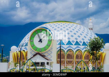 palu, Indonesien - 10. März 2026 : Vorderfassade der Baitul-Khairaat-Moschee mit einer massiven analogen Uhr, eingebettet in eine rautengemusterte Kuppel, großen bogenförmigen gelben Fenstern und einem bergigen Hintergrund an einem bewölkten Tag. Stockfoto