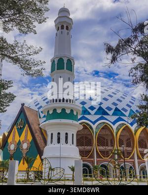 Palu, Indonesien - 10. März 2026 : lebendige Aufnahme eines weißen Minaretts in der Baitul Khairaat Grand Mosque, vor einem hellblauen Himmel mit wackeligen Wolken. Das Foto zeigt auch die komplizierten Details der Kuppel und die gelben Zierfenster. Stockfoto