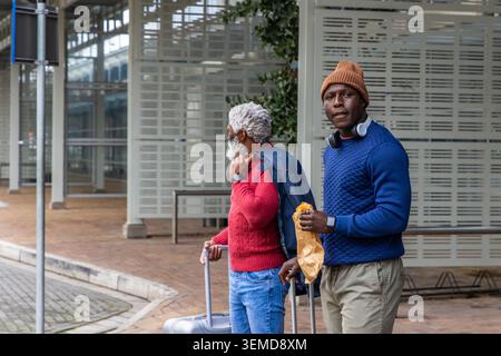 Zwei Männer in rotem Strick und brauner Mütze, ziehen Koffer an der Station und halten Papiertüte Stockfoto