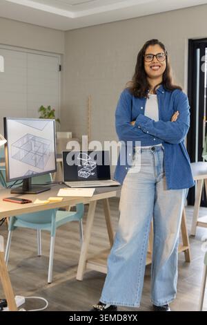 Asiatische Frau Ende 20, die mit gekreuzten Armen im Studio steht und CAD auf Monitor und Laptop zeigt Stockfoto