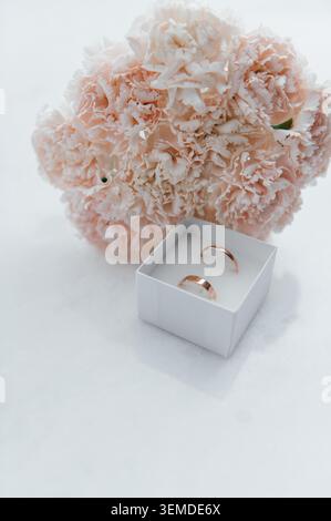 Zwei Eheringe aus Roségold in einer weißen Geschenkbox mit einem Blumenstrauß aus zarten rosa Nelken. Stockfoto