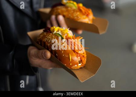 Nahaufnahme einer Person, die einen Streetfood-Hotdog in einem Sesambrötchen hält, garniert mit Karottenkrautsalat, Gurke und Senf, bereit zum Verzehr als schnelle Take-away s Stockfoto