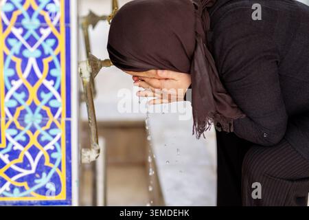 Muslimische Frau im Hijab, die ihr Gesicht mit fließendem Wasser an einem Moschee-Brunnen wäscht und vor dem Gebet wudu vorführt, neben farbenfrohen islamischen Fliesenmustern. Stockfoto