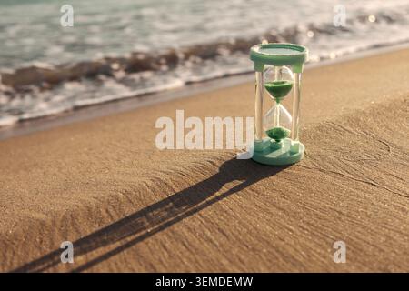 Eine Sanduhr mit grünem Sand liegt an einem sonnigen Sandstrand nahe dem Meer und wirft einen langen Schatten. Der Hintergrund zeigt sanfte Wellen, die sich t nähern Stockfoto