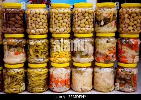 Nahaufnahme von gestapelten Plastikgläsern gefüllt mit Oliven, Paprika und gemischtem eingelegtem Gemüse an einem Marktstand, auf dem bunte Konserven und gezeigt werden Stockfoto